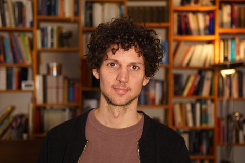 Porträt einer Person mit lockigem dunklem Haar vor einem Bücherregal.