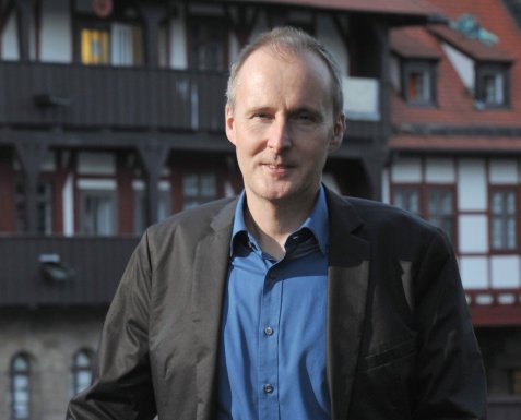 Mann mit blauem Hemd und Sakko steht lächelnd vor einem Fachwerkhaus.