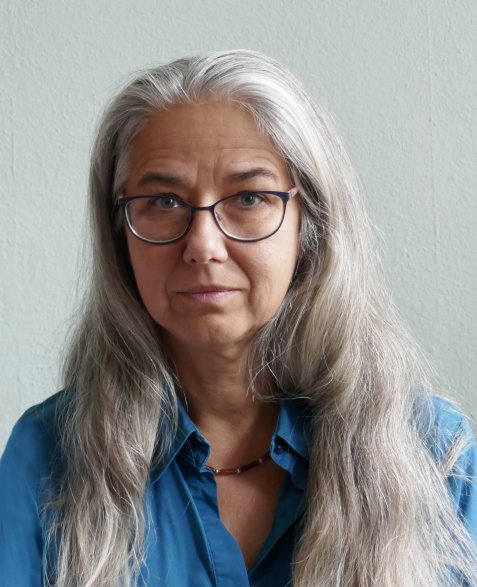 Porträt einer Person mit langen grauen Haaren, Brille und blauer Bluse vor heller Wand.