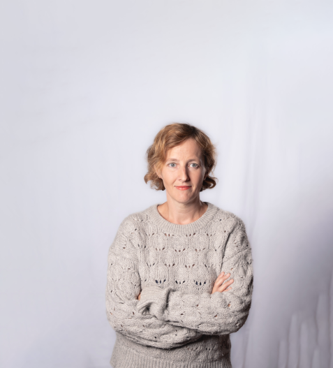 Porträt einer Frau mit kurzen, lockigen Haaren und beigem Strickpullover, die mit verschränkten Armen vor hellem Hintergrund steht.