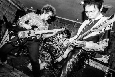 Schwarz-weißes Bühnenfoto einer Band mit zwei Gitarristen im Vordergrund und einem Schlagzeuger im Hintergrund während eines energiegeladenen Live-Auftritts.