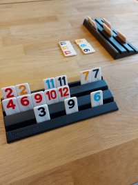 Spielsteine mit Zahlen auf einem Rummikub-Spielhalter sowie weitere Steine und ein leerer Halter auf einem Holztisch.