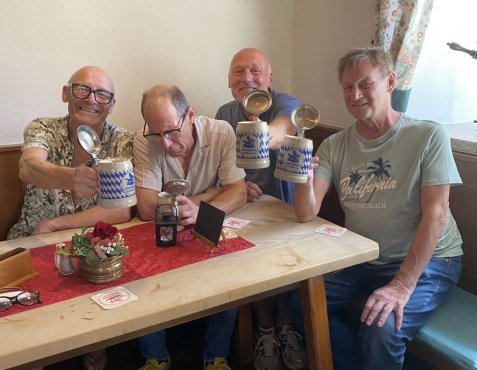 Vier lächelnde Männer sitzen an einem Tisch in einer Gaststube und heben ihre Bierkrüge mit Deckel zum Anstoßen.