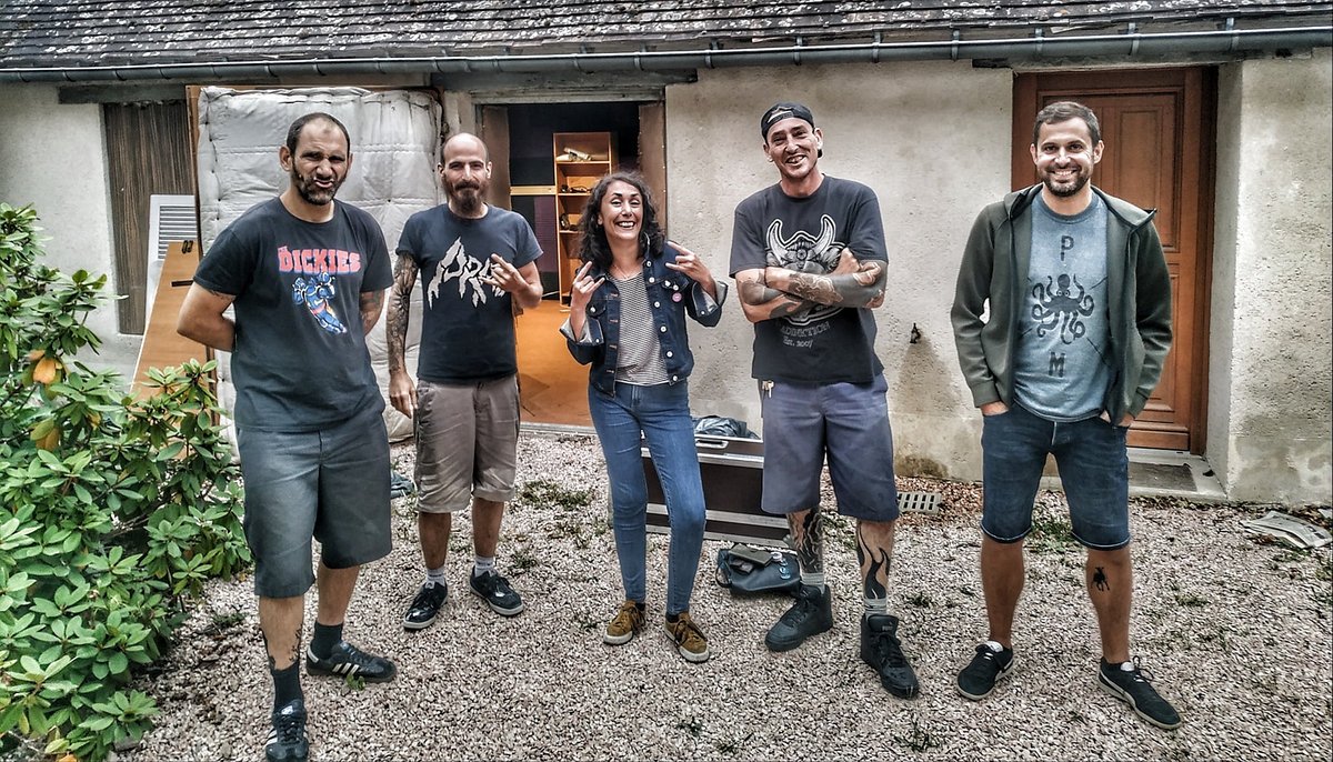 Fünf lachende Personen stehen in kurzen Hosen und T-Shirts vor einem Haus mit offener Garage im Hof.