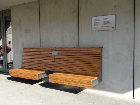 Holzbank aus Latten vor dem Gebäude des Wohnprojektes Spiegelfabrik. An der Wand ist eine Gedenktafel zur Erinnerung an die enteignete jüdische Familie Midas angebracht.