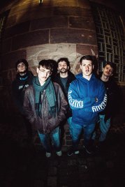 Fünf Personen in Alltagskleidung stehen mit ernstem Blick vor einer alten Steinmauer, aufgenommen mit einem Fisheye-Objektiv.