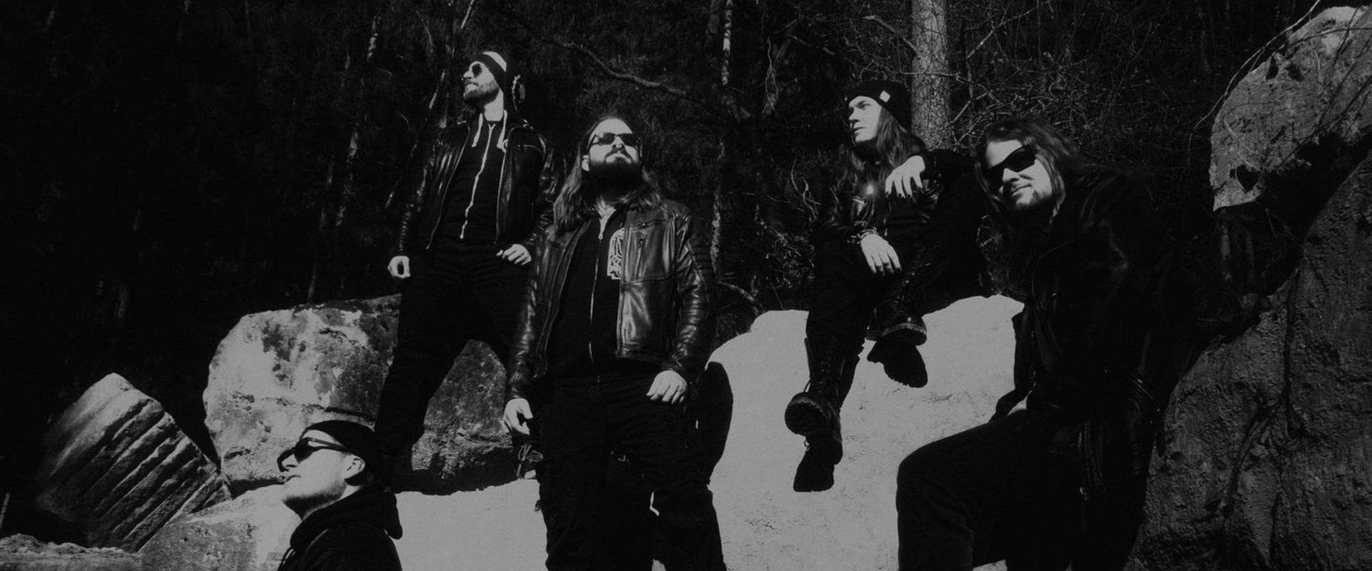 Schwarz-weißes Bandfoto von fünf Personen in schwarzer Kleidung und Sonnenbrillen, die auf Felsen in einem Wald posieren.