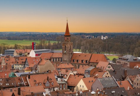 Blick über die Altstadt mit roten Ziegeldächern und einem hohen Kirchturm mit spitzem Dach vor abendlichem Himmel.