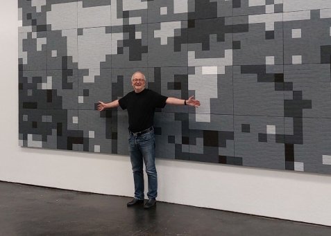 Eine Person breitet lächelnd die Arme vor einem großformatigen Kunstwerk mit grau-schwarzem Pixelmuster aus.