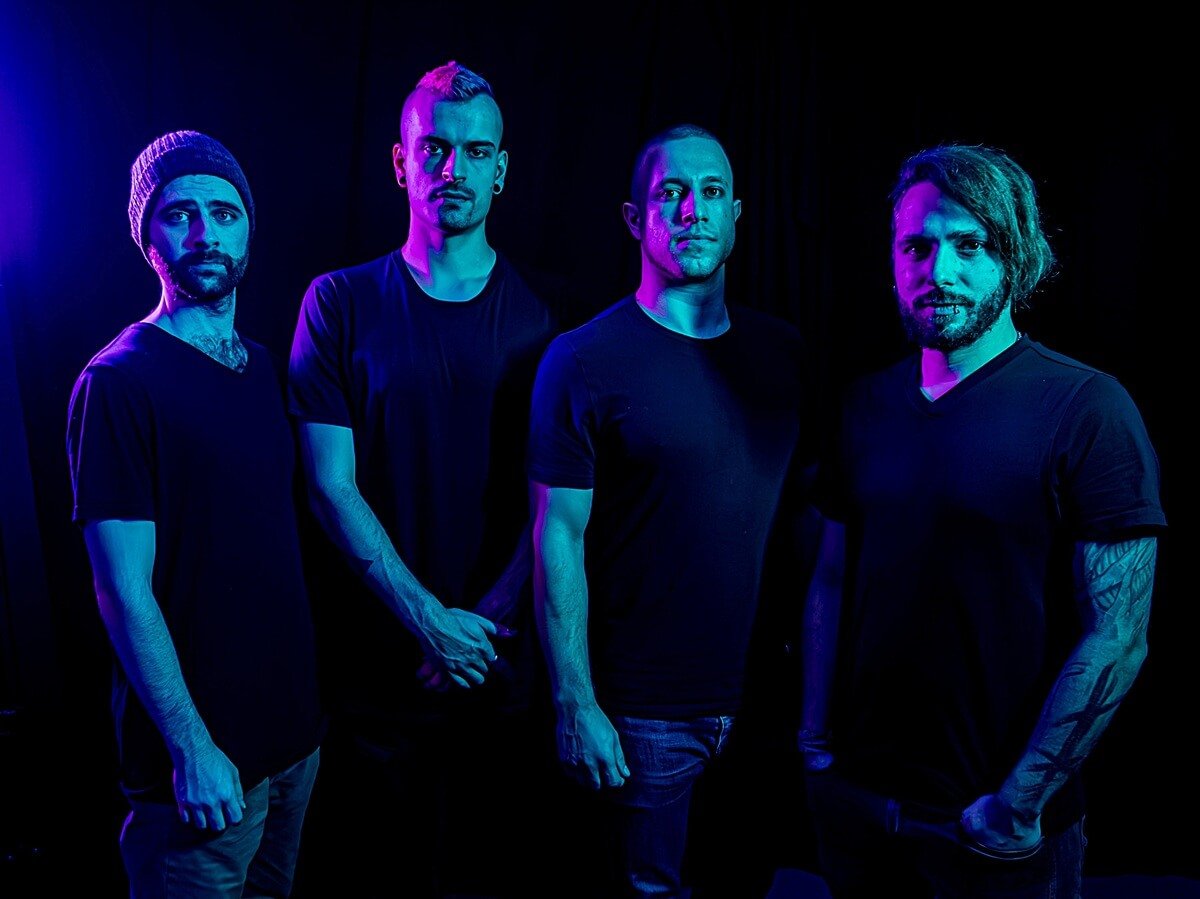 Vier Personen in dunklen T-Shirts stehen nebeneinander vor schwarzem Hintergrund, in blaues und violettes Licht getaucht.