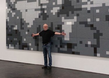 Eine Person breitet lächelnd die Arme vor einem großformatigen Kunstwerk mit grau-schwarzem Pixelmuster aus.