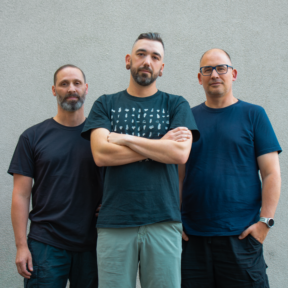 Drei Personen in T-Shirts stehen nebeneinander vor einer grauen Wand und blicken in die Kamera.