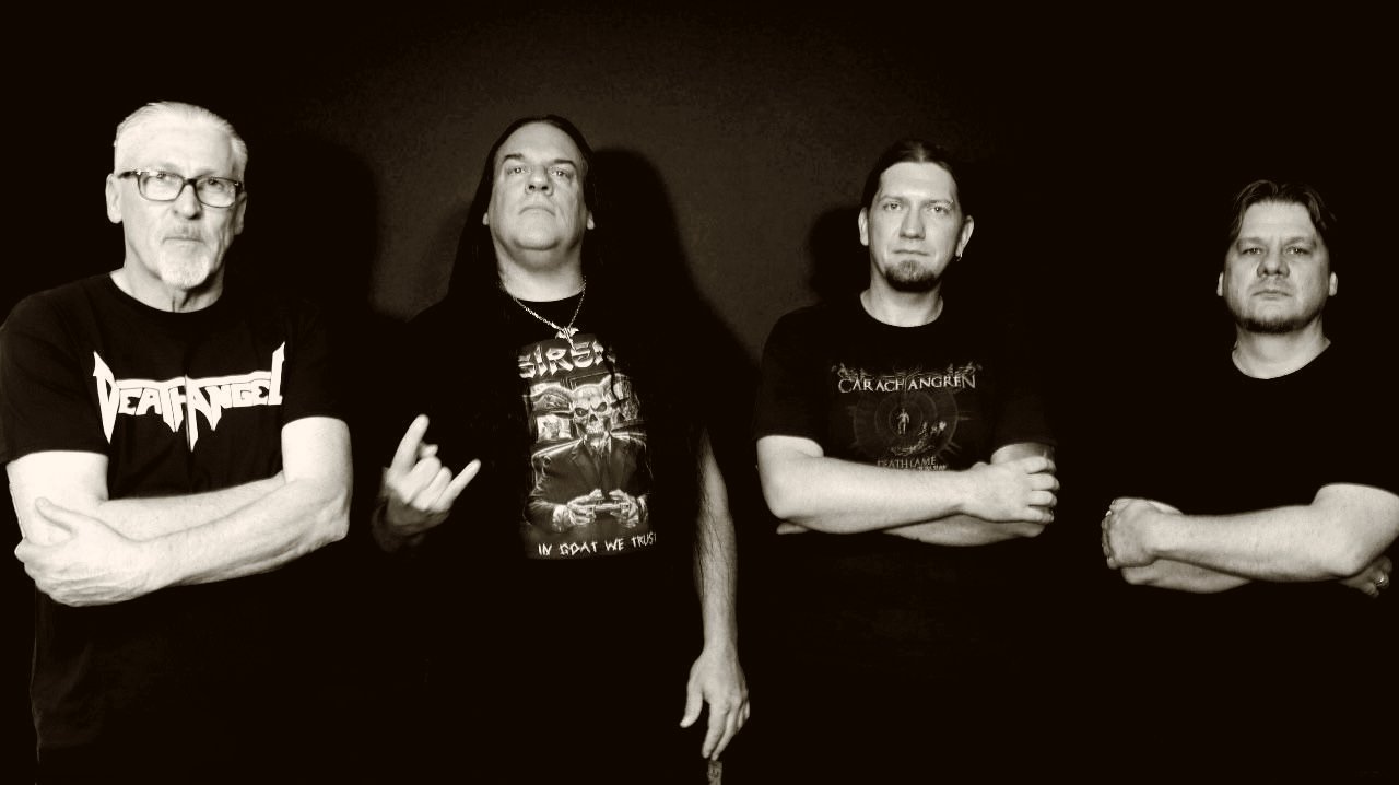 Vier Männer in Bandshirts posieren mit verschränkten Armen vor dunklem Hintergrund, einer zeigt das Metal-Horns-Zeichen.