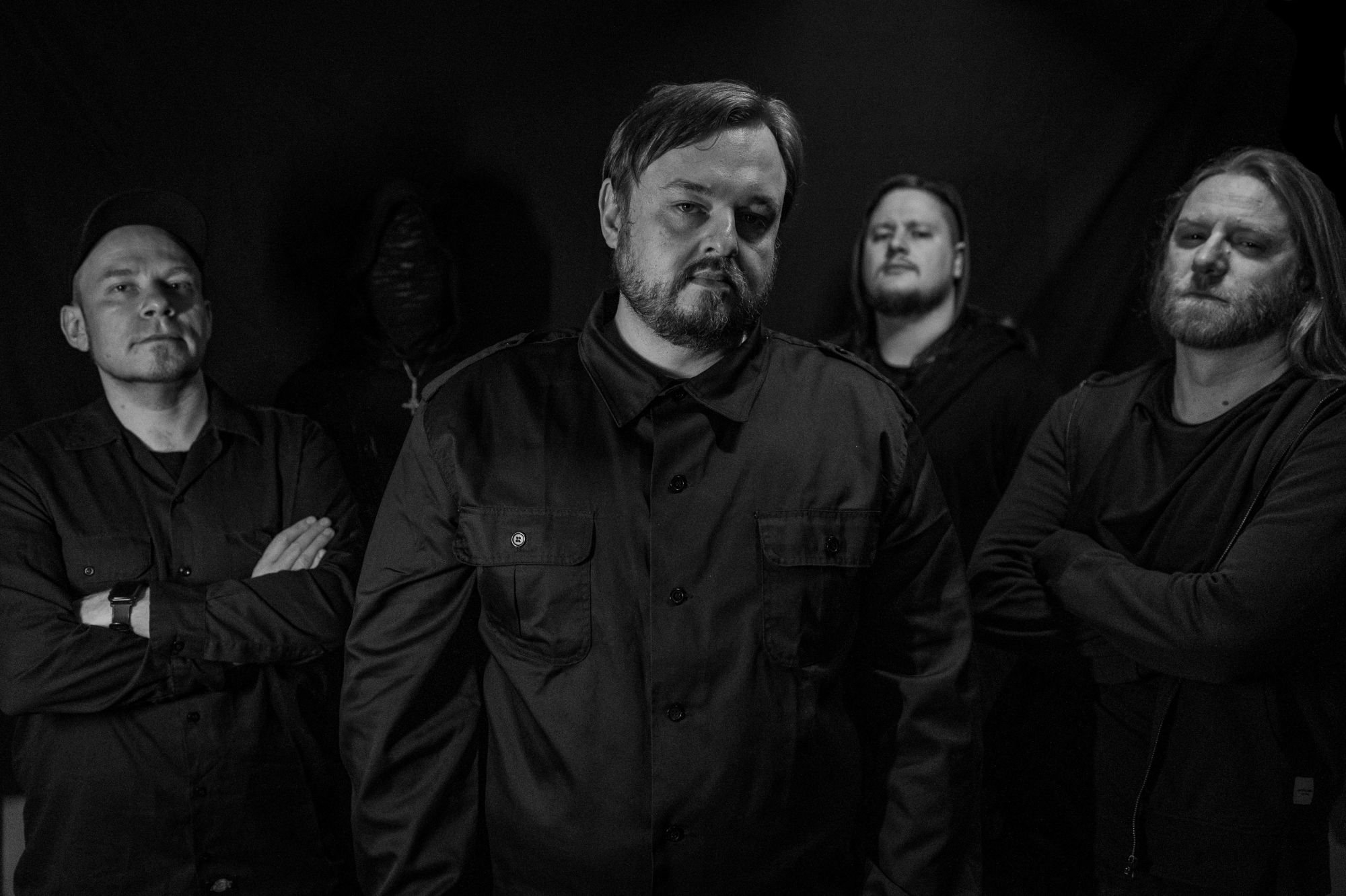 Schwarz-Weiß-Porträt einer fünfköpfigen Band in dunkler Kleidung, im Hintergrund steht eine vermummte Person mit Kapuze.