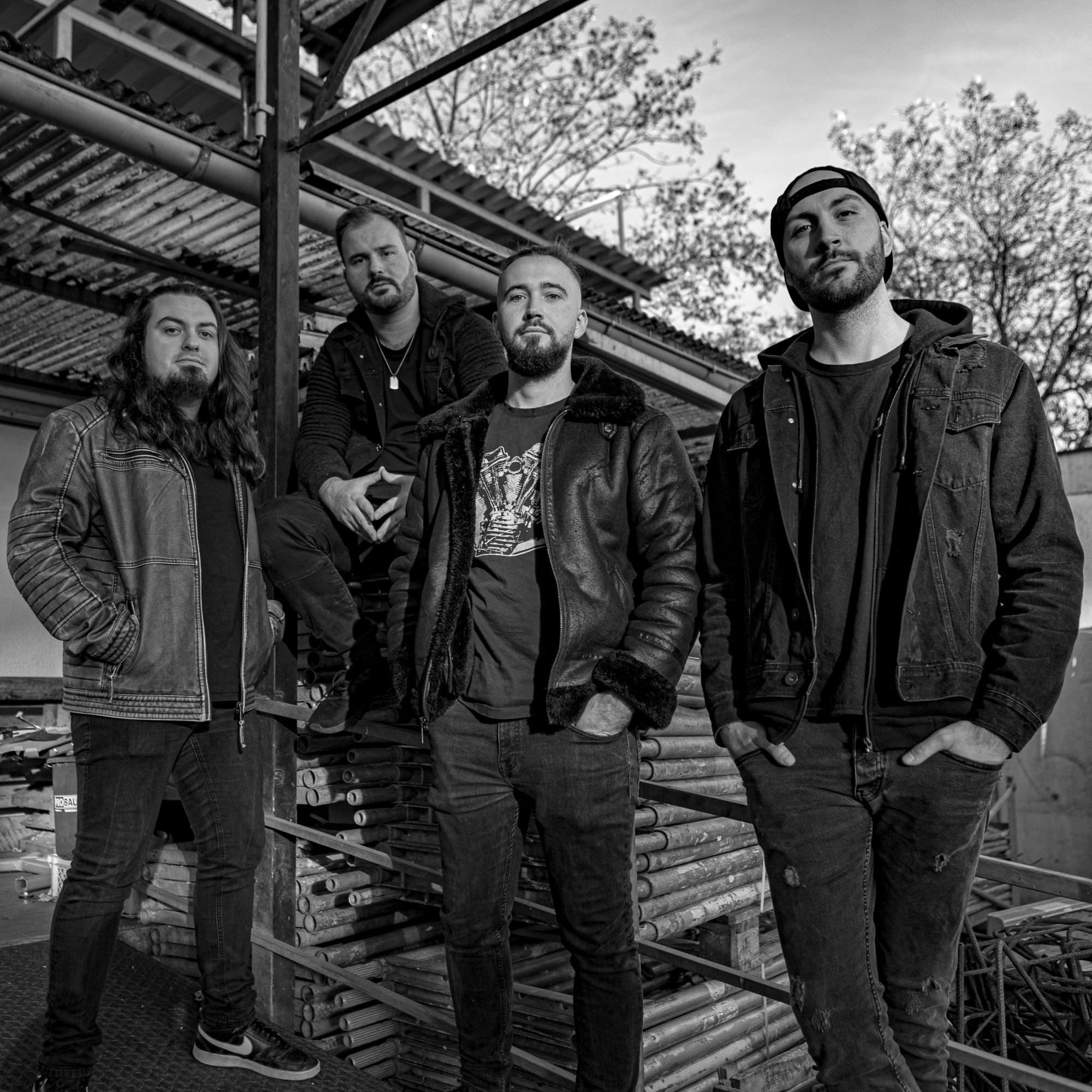 Schwarz-weißes Bandfoto von vier Männern in Lederjacken, die zwischen Baugerüst und gestapelten Metallrohren stehen und in die Kamera blicken.