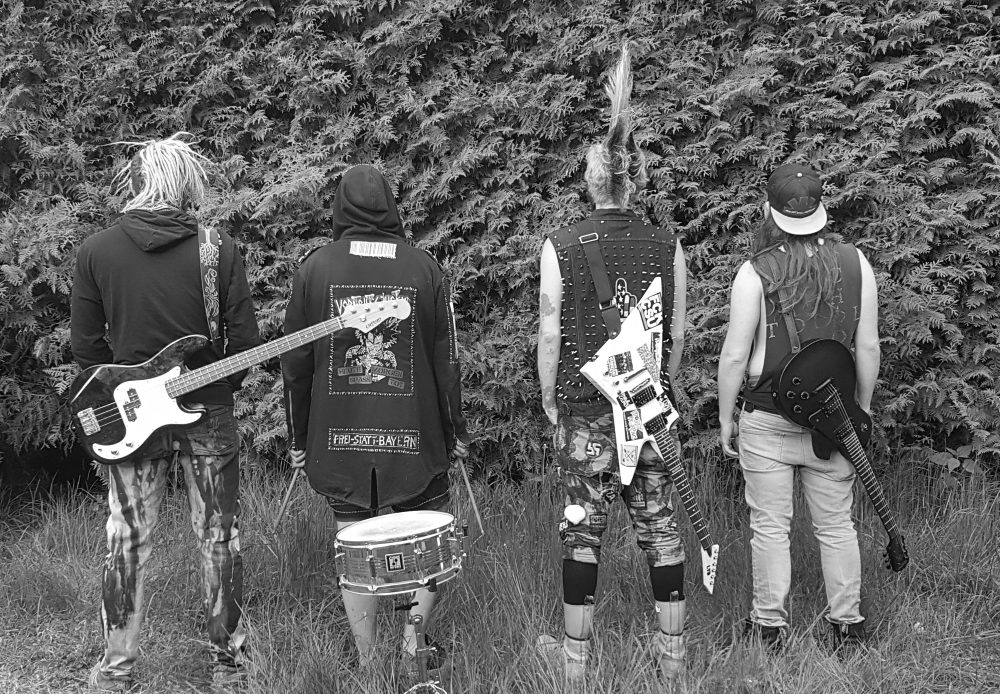 Vier Punk-Musiker mit Gitarren und einer Snaredrum stehen von hinten fotografiert vor einer dichten Hecke.