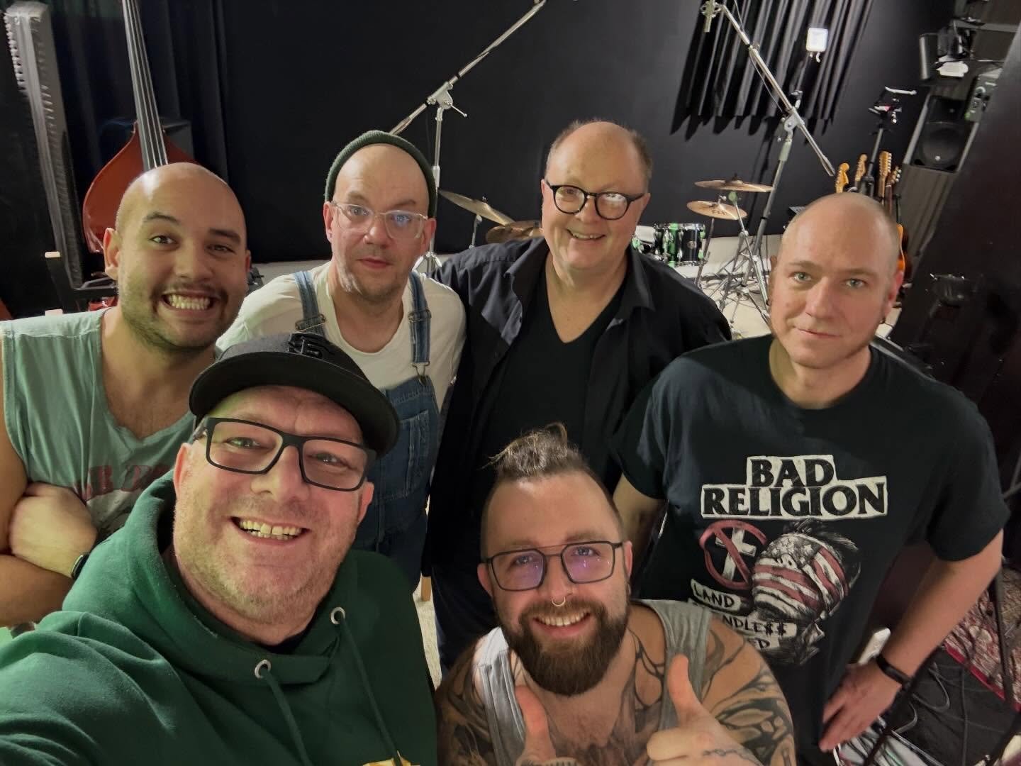 Sechs lächelnde Personen posieren für ein Gruppen-Selfie in einem Musikproberaum mit Kontrabass, Schlagzeug und Mikrofonständern im Hintergrund.