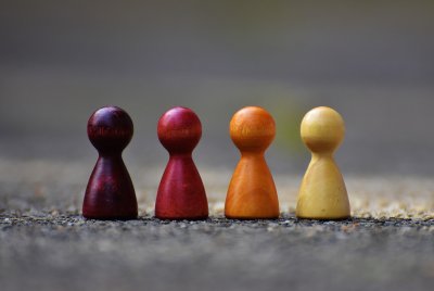 Vier hölzerne Spielfiguren in unterschiedlichen Farbtönen von dunkelviolett über rot und orange bis gelb stehen nebeneinander auf einer steinernen Oberfläche.