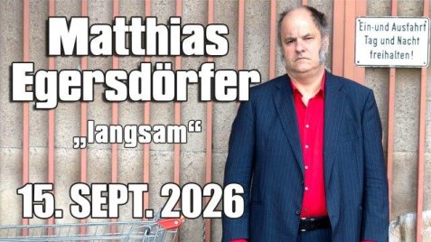 Matthias Egersdörfer mit rotem Hemd und dunklem Sakko steht neben einem Einkaufswagen vor einer Wand mit senkrechten Streifen, daneben der Text 'Matthias Egersdörfer "langsam" 15. SEPT. 2026'