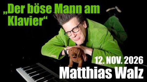 Matthias Walz mit schwarzer Brille und grünem Sakko liegt auf dem Boden neben einem Klavier und hält ein braunes Stoffhundspielzeug