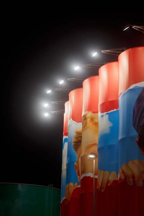 Große zylindrische Silos mit einem großflächigen Wandbild eines Kindes werden nachts von Scheinwerfern angestrahlt.