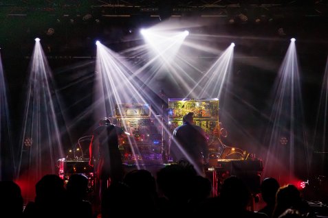 Zwei Musiker stehen auf einer Bühne an modularen Synthesizer-Anlagen, während Scheinwerfer durch Nebel strahlen und das Publikum davor im Dunkeln steht.