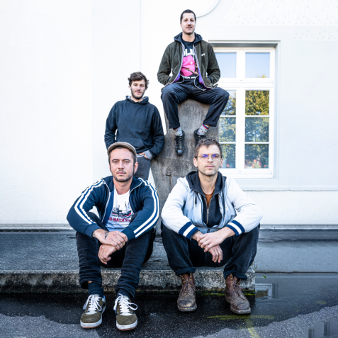 Vier Personen posieren an einer steinernen Sockelkonstruktion, zwei sitzen unten, zwei stehen/sitzen erhöht dahinter.