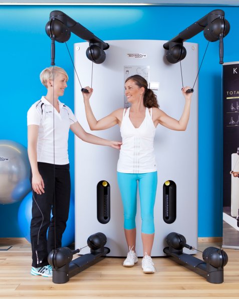 Eine Trainerin betreut eine Frau beim Training an einem Kabelzug-Fitnessgerät der Marke TechnoGym.