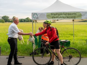 OB Jung überreicht im Rahmen des Pendlerfrühstücks einer Radfahrerin ein kleines Stoffbeutel-Geschenk an einem Info-Pavillon des Stadtplanungsamts Fürth an einem Radweg.