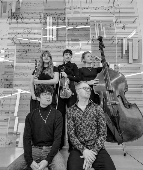 Fünf Musikerinnen und Musiker mit Klarinette, Violine und Kontrabass posieren in Schwarz-Weiß vor einer Glaswand mit handgeschriebenen Notenblättern.