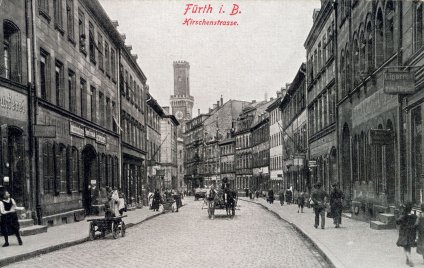 Historische Schwarz-Weiß-Aufnahme der Hirschenstraße in Fürth mit Kopfsteinpflaster, Kutschen und Fußgängern zwischen mehrstöckigen Häuserzeilen, im Hintergrund ein Turm.