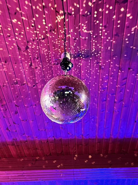 Eine glitzernde Discokugel hängt von einer Holzdecke und wirft unter violett-pinker Beleuchtung viele kleine Lichtreflexe an die Decke.