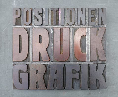 Drei Reihen mit großen Metalllettern in Großbuchstaben, die die Wörter 'POSITIONEN', 'DRUCK' und 'GRAFIK' bilden.