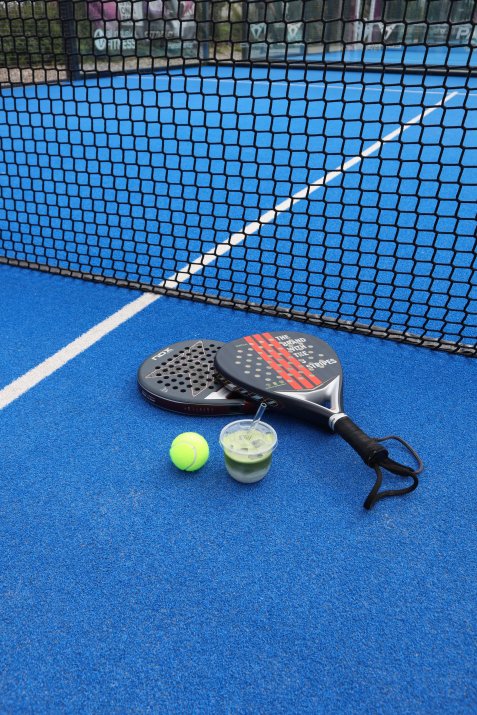 Zwei Padelschläger, ein Padel-Ball und ein Becher gefüllt mit einem Iced Matcha Latte vor dem Netz eines Padelplatzes.