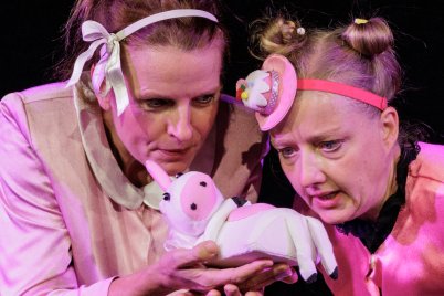 Zwei Schauspielerinnen in rosafarbenen Kostümen betrachten bei einer Theateraufführung gemeinsam ein kleines Stofftier in Form eines weißen Einhorns.