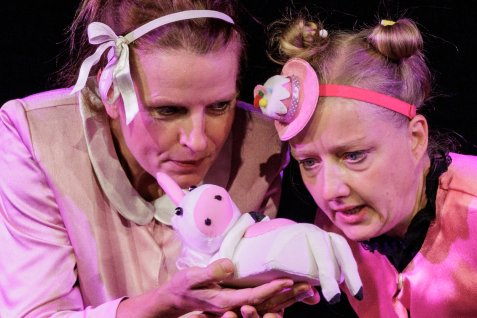 Zwei Schauspielerinnen in rosafarbenen Kostümen betrachten bei einer Theateraufführung gemeinsam ein kleines Stofftier in Form eines weißen Einhorns.
