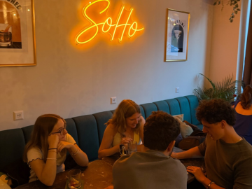 Mehrere Personen sitzen lachend an einem Tisch auf einer blauen Sitzbank unter einem leuchtenden „SoHo“-Schriftzug.