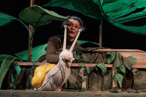 Schauspielerin führt auf einer Theaterbühne eine große Schneckenpuppe mit gelbem Haus vor einer Kulisse aus grünen Blättern.