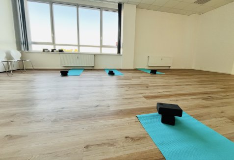 Heller Yogaraum mit türkisfarbenen Yogamatten und schwarzen Yogablöcken auf Holzboden vor einer großen Fensterfront.