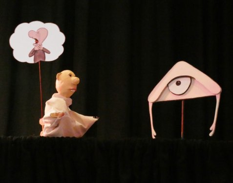Eine Puppenfigur mit Gedankenblase steht auf einer Bühne neben einer dreieckigen Figur mit großem Auge und dünnen Beinen vor schwarzem Vorhang.