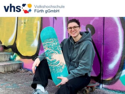 Eine lächelnde Person mit Brille sitzt vor einer bunten Graffiti-Wand und hält ein türkisfarben bemaltes Skateboard, oben das Logo der Volkshochschule Fürth gGmbH.