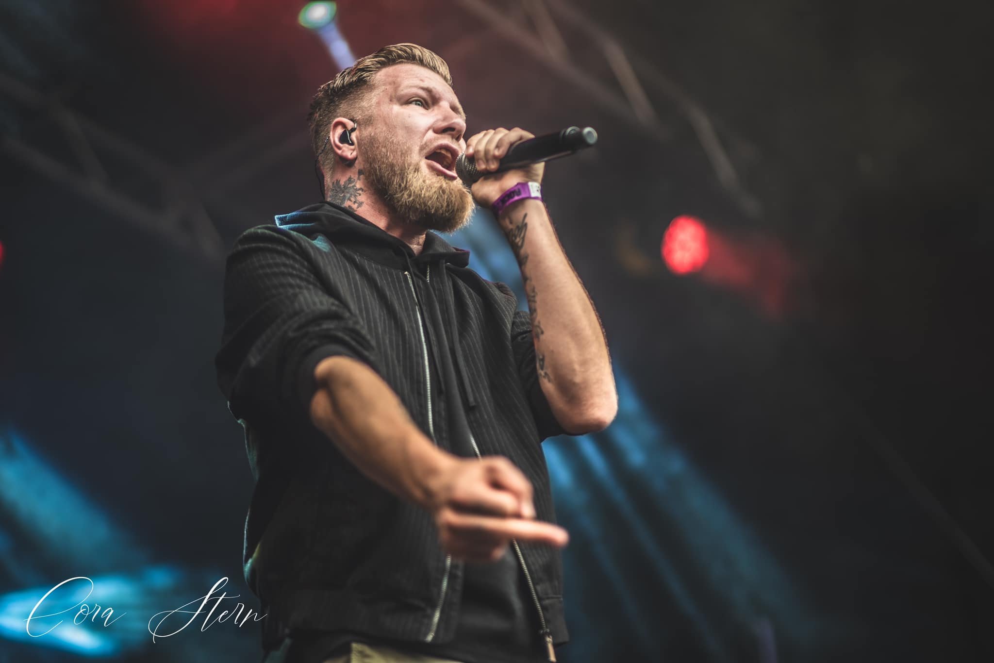 Bärtiger Sänger mit Mikrofon und In-Ear-Monitoren performt auf einer Bühne mit blauer und roter Beleuchtung.