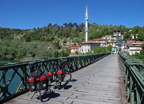 Ein mit Gepäcktaschen beladenes Fahrrad steht auf einer grünen Stahlbrücke, im Hintergrund sind ein Minarett und eine Siedlung am Fluss zu sehen.