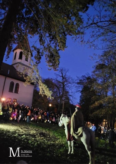 Person in Mantel und mit rotem Helmbusch auf einem Schimmel vor einer Menschenmenge mit Laternen an einer Kirche in der Abenddämmerung
