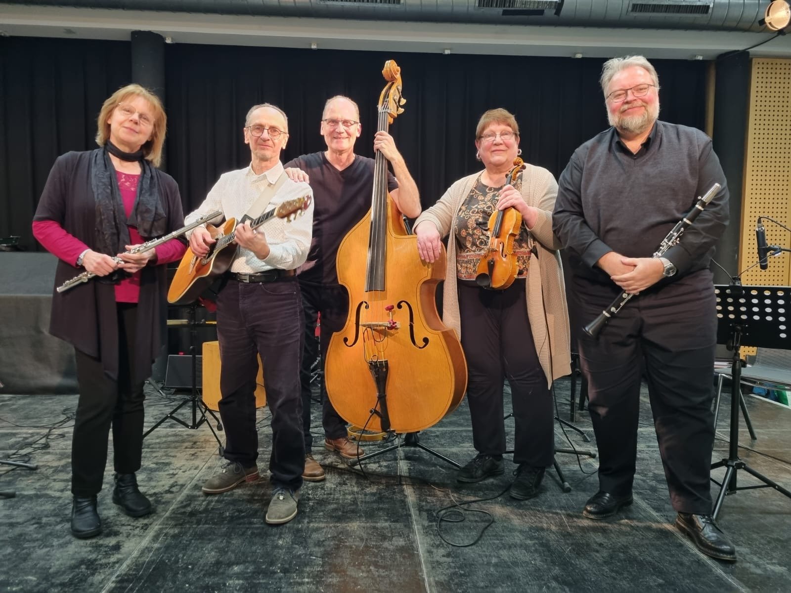 Fünf Musizierende stehen lächelnd nebeneinander auf einer Bühne und halten Querflöte, Gitarre, Kontrabass, Geige und Klarinette in den Händen.