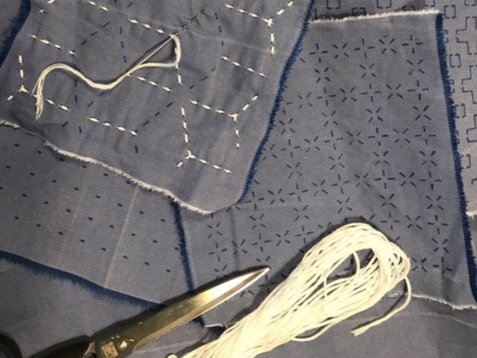 Blaue Stoffstücke mit vorgezeichneten Mustern und weißen Stichen in Sashiko-Art, daneben liegen eine Schere und ein Bündel weißes Stickgarn.