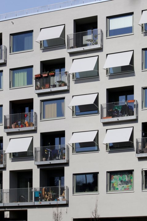 Wohnhausfassade mit Fenstern, ausgestellten hellen Markisen und kleinen Balkonen, auf denen Pflanzen, Stühle und Alltagsgegenstände stehen.