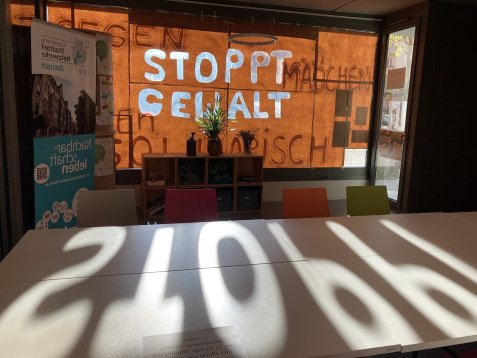 Blick aus dem Quartiersbüro auf das orange beklebte Schaufenster mit der Aufschrift „Stoppt Gewalt“, davor ein Tisch mit dem Schatten der Buchstaben.